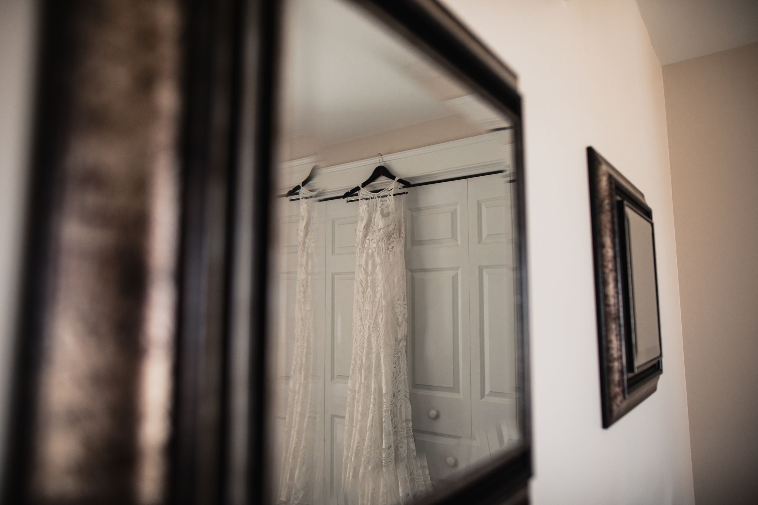 A delicate lace wedding dress hanging on a hanger, reflected in an ornate dark-framed mirror in a bridal suite.