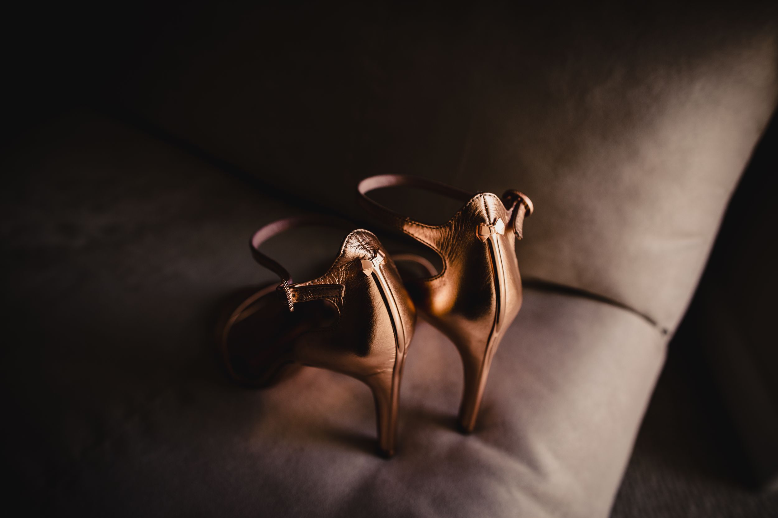 Elegant rose gold strappy high heel sandals resting on a gray velvet sofa in soft light.