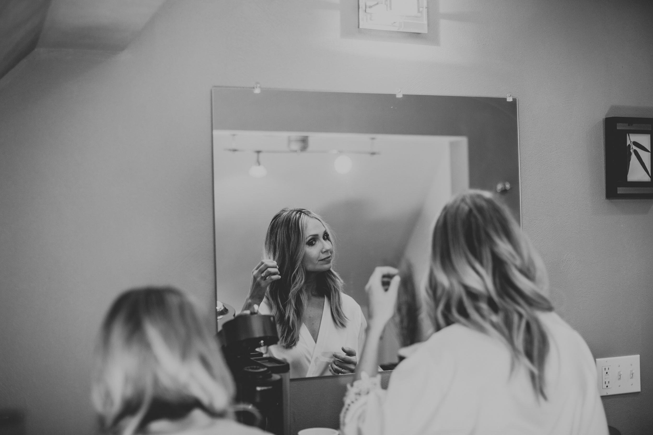 Black and white photo of a bride in a white robe applying makeup in front of a mirror while getting ready.