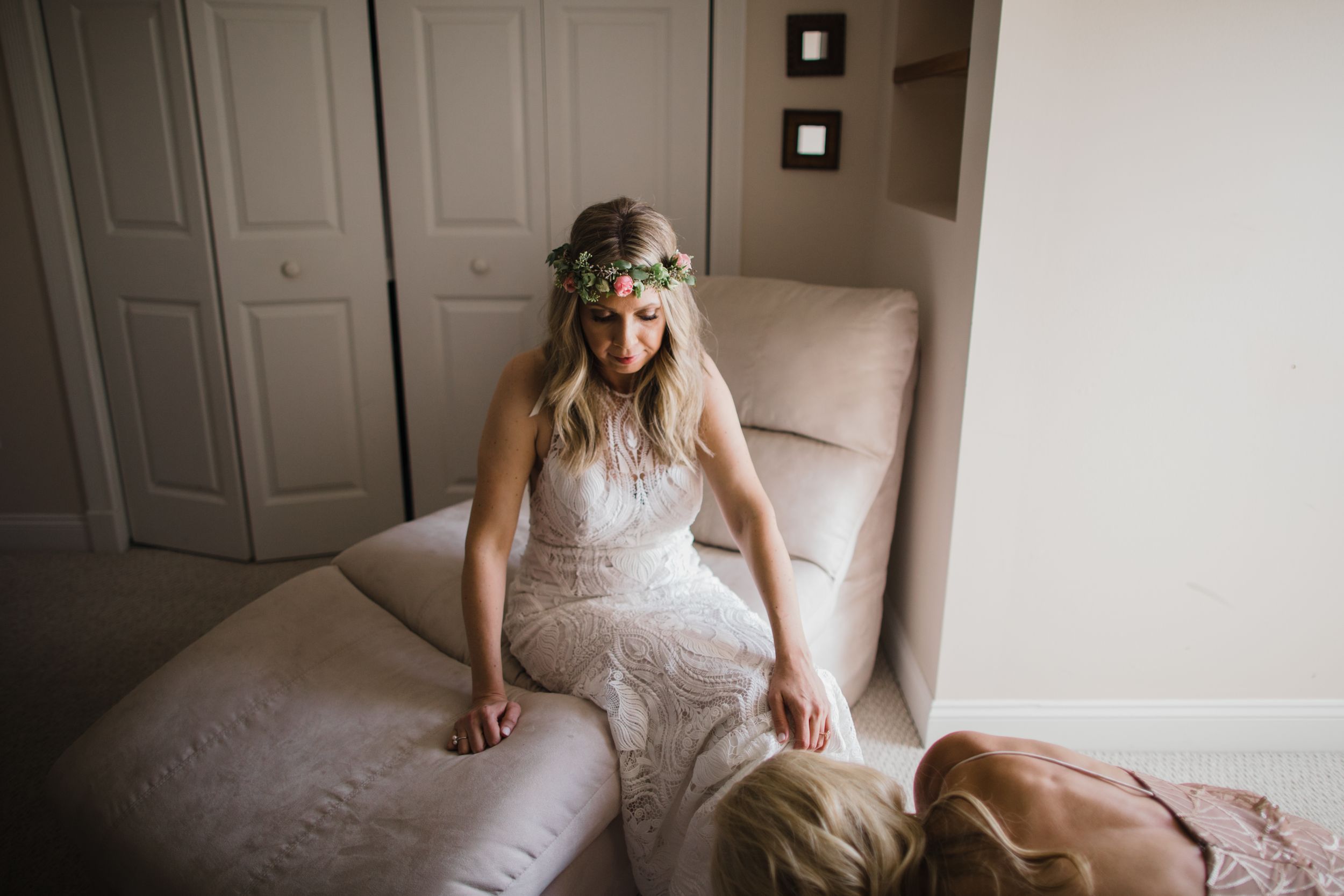 Bride in lace wedding dress wearing a floral crown sits on a cream chaise lounge before ceremony.