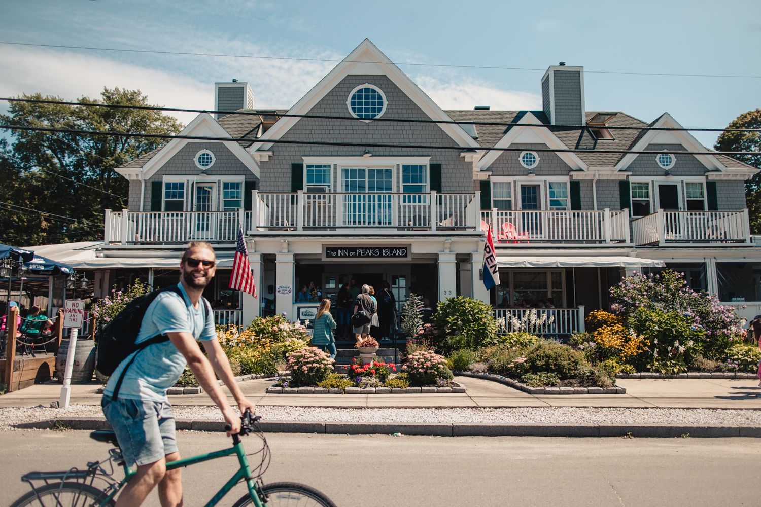 Man riding a bike past a charming New England inn adorned with flowers and American flags on a sunny day.