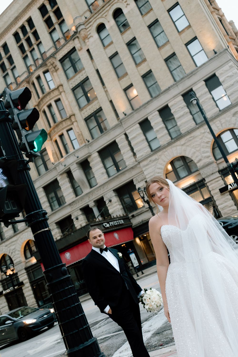Editorial Bride and groom posing on a city street corner near The Pfister Hotel, bride in strapless gown with veil, groom in black tuxedo.