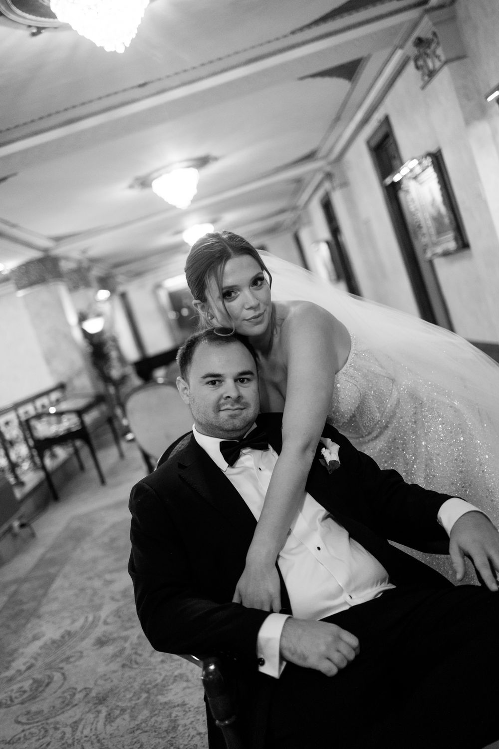 Black and white editorial wedding photo of bride leaning over seated groom in tuxedo, posing elegantly in ornate venue.