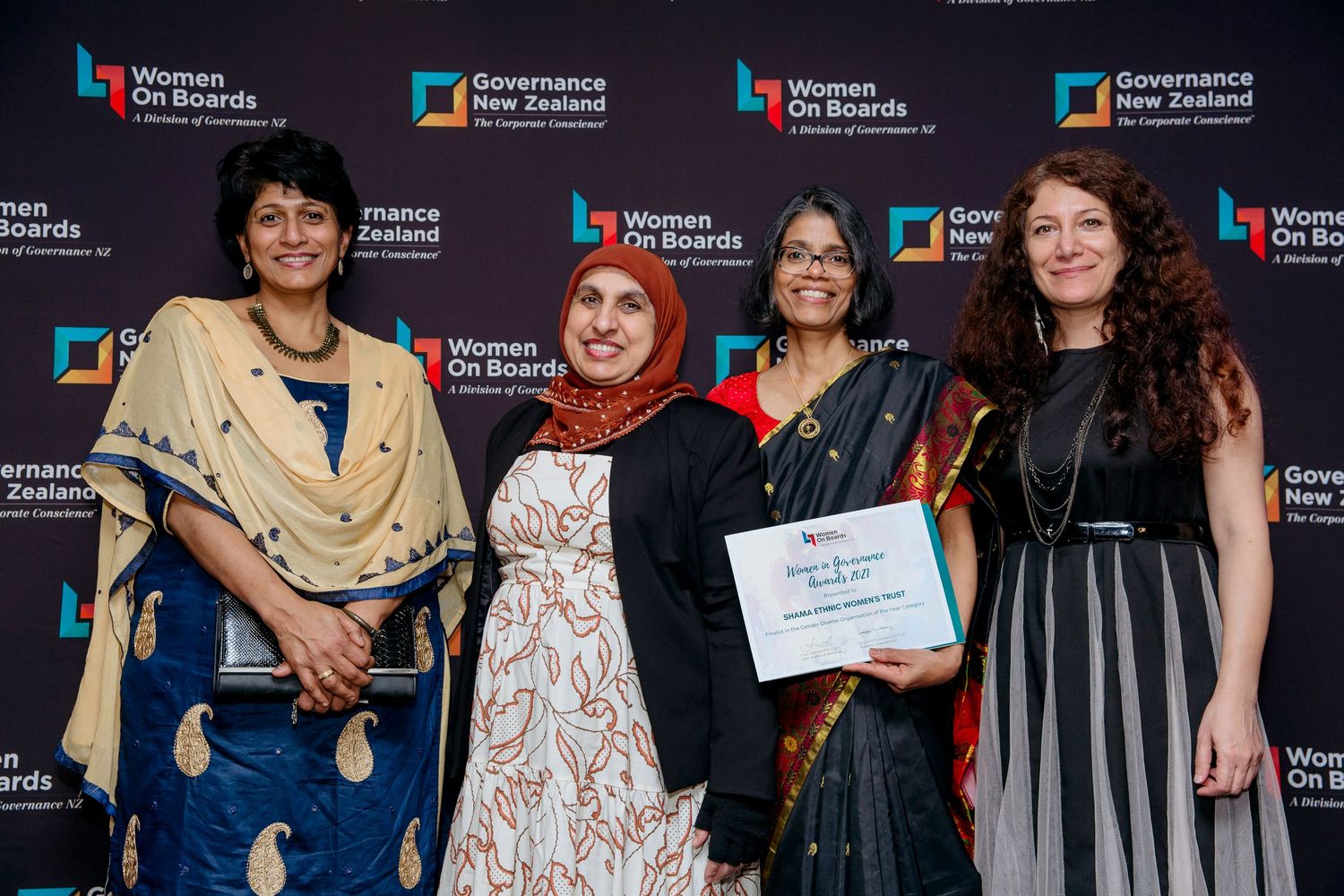 Four women at a corporate event standing in front of a media wall with the certificate they were awarded