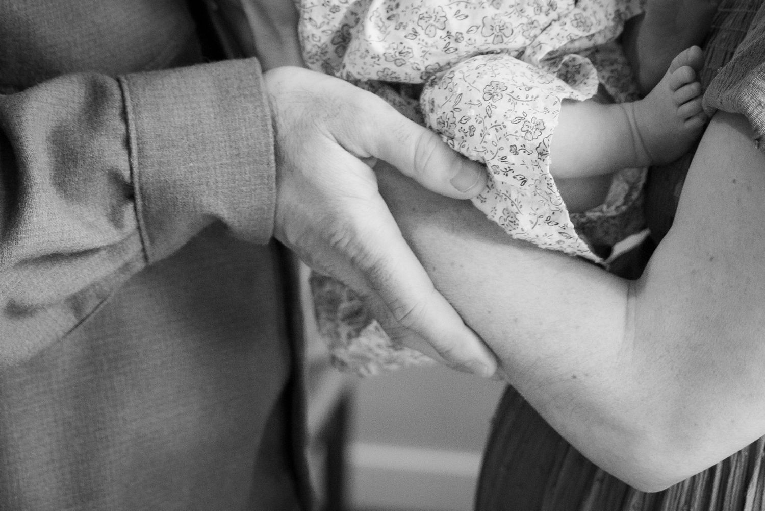 Black and white close-up of father holding newborn baby in floral outfit, hands intertwined.