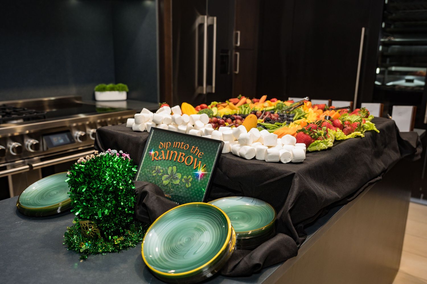 St. Patricks Day party food spread with colorful fruit platter and green decorative plates on a dark table.