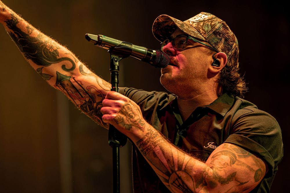 A performer in a dark cap sings passionately into a microphone during a live concert performance.