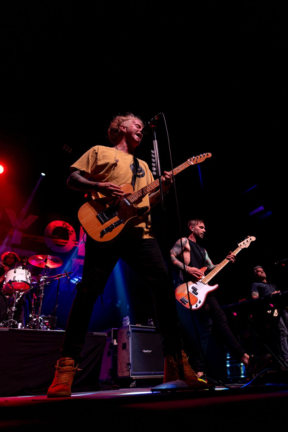 Musicians perform energetically on stage with guitars under dramatic red and blue concert lighting.