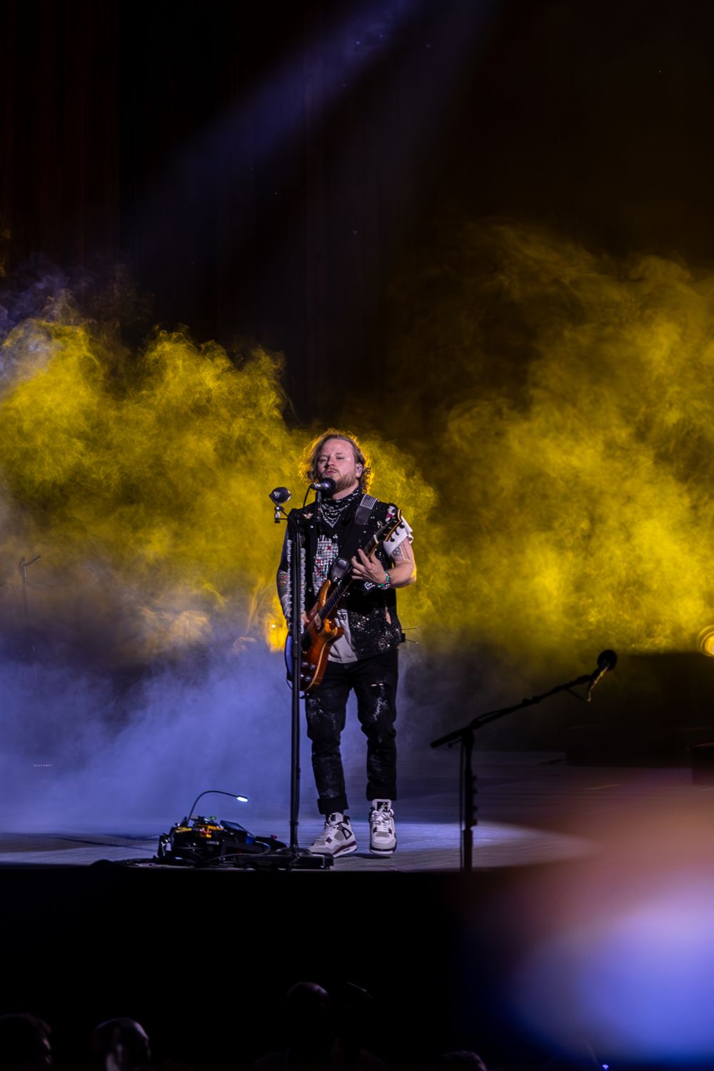 A performer stands on stage surrounded by dramatic yellow smoke effects and blue lighting during a concert performance.