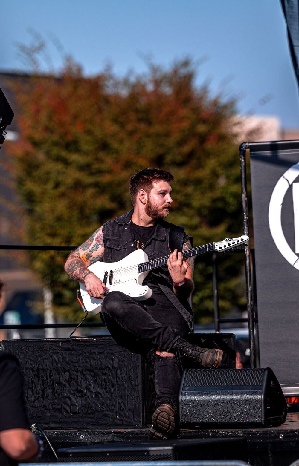 Rock musician performs on stage with white electric guitar against outdoor backdrop during daytime concert.