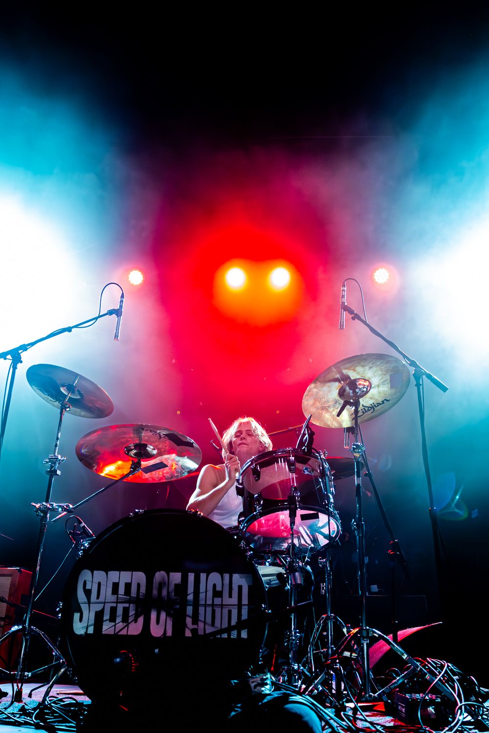 Concert stage setup with drums and equipment illuminated by dramatic red and blue lighting effects during performance.