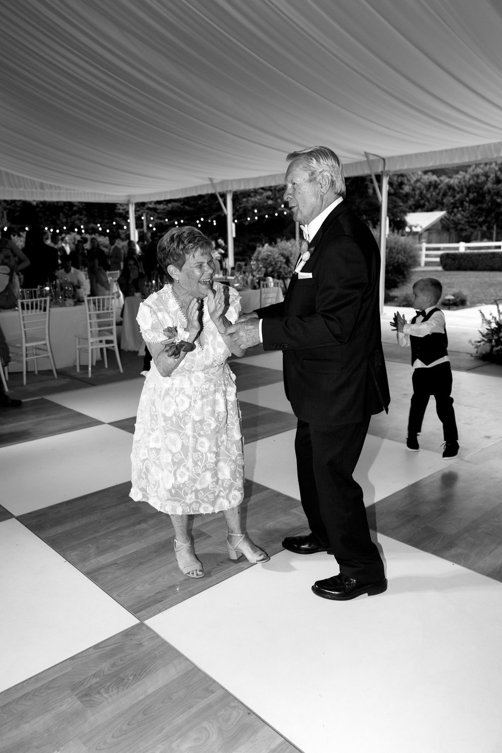 A couple dances together on a black and white checkered dance floor under an elegant tent at a formal event.