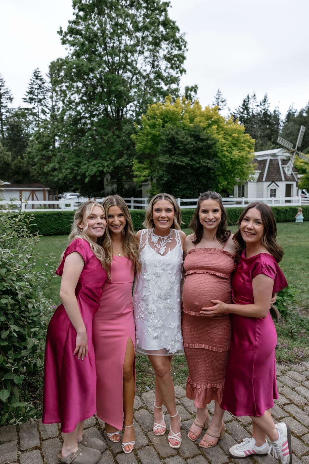 A group in coordinating pink and burgundy dresses pose together outdoors in a garden setting.