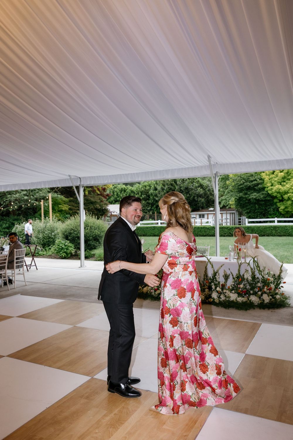 A couple dances together under a white tent on a checkered dance floor at an outdoor wedding reception.