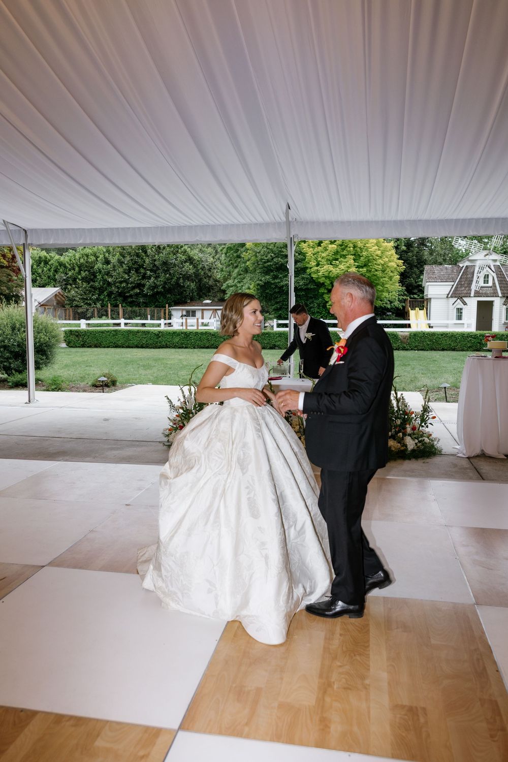 A couple in formal wedding attire dances together under an elegant white tent on a wooden dance floor.