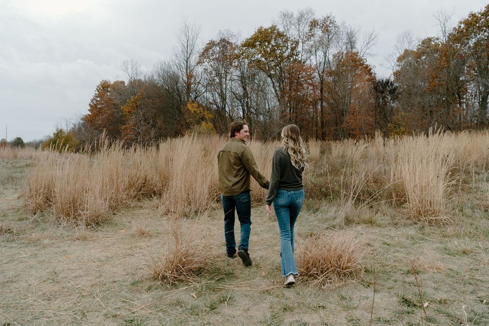 Two people walk through a tall grass meadow with autumn trees in the background on a cloudy day.