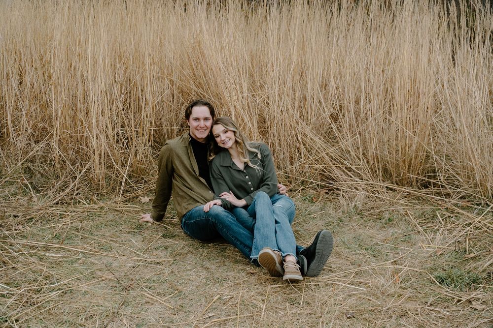 A couple sits together in tall brown grass on a cloudy autumn day wearing casual clothing.