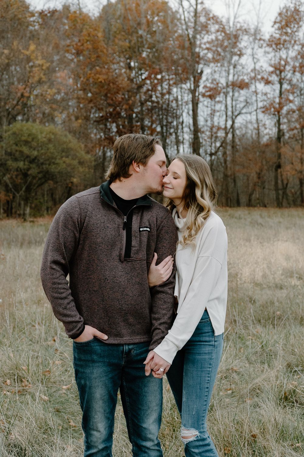 A couple shares tender moments in a field surrounded by autumn trees and tall grass on an overcast day.