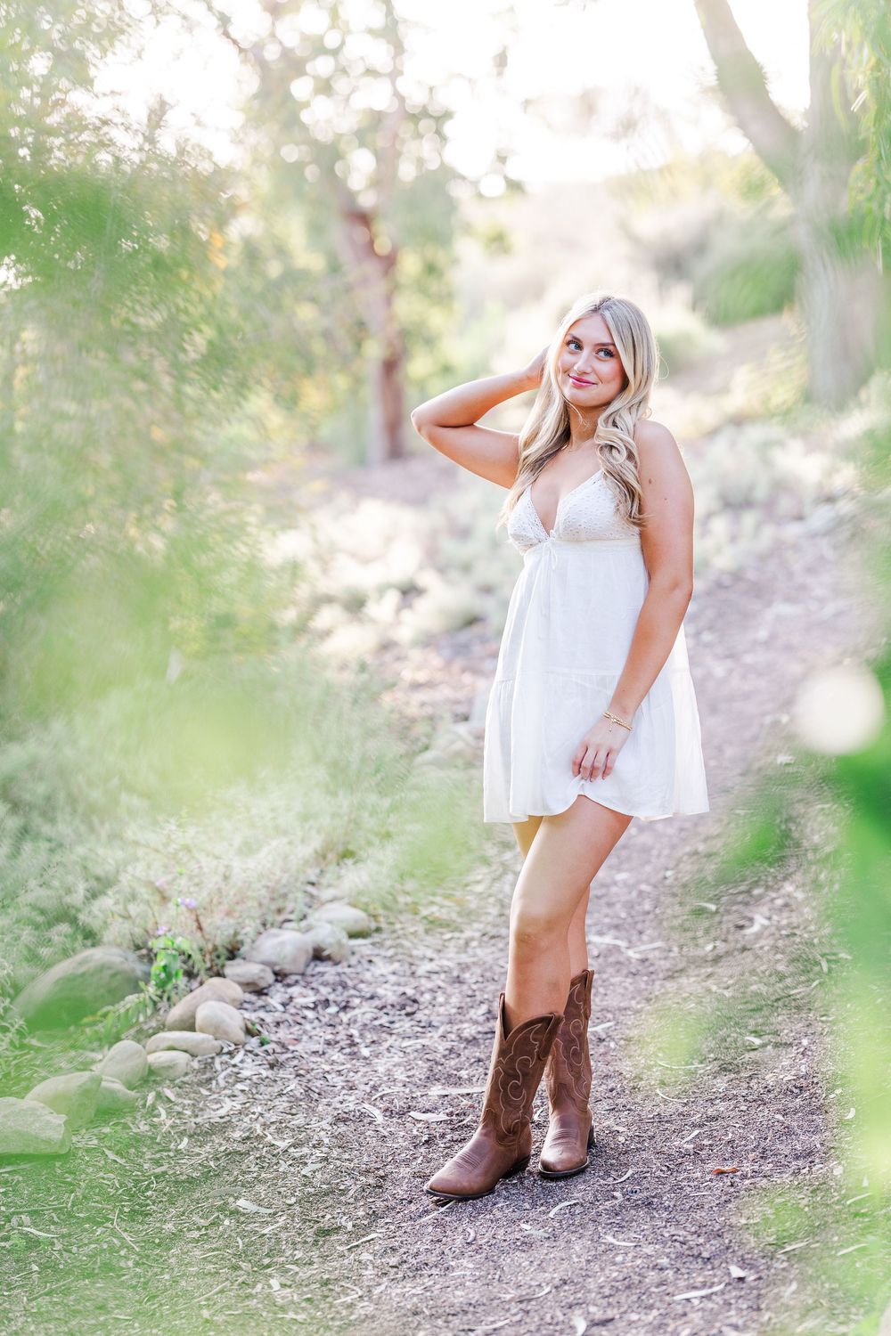 A woman in a white dress and brown cowboy boots poses on a rocky trail surrounded by greenery.