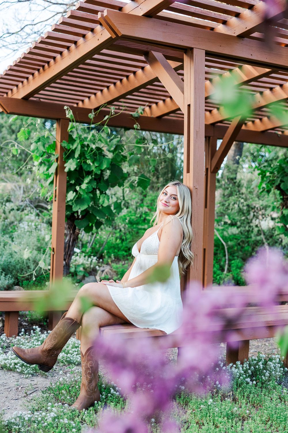 A white dress contrasts beautifully against purple flowers and a wooden pergola in an outdoor garden photoshoot.