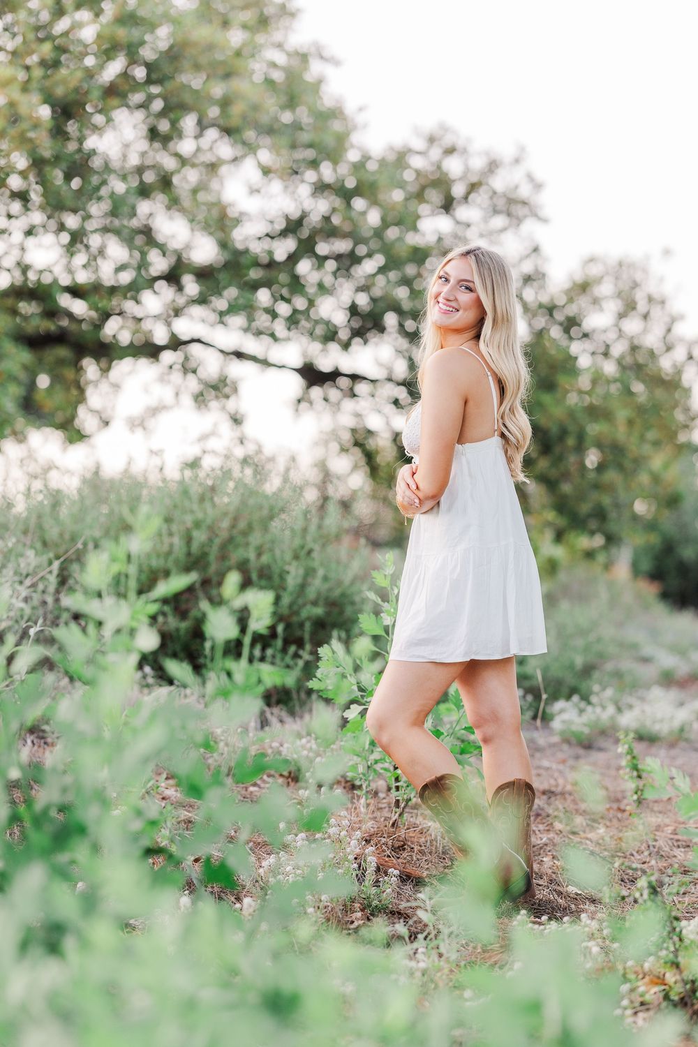 A white sundress and boots outfit is showcased against a lush green natural backdrop.