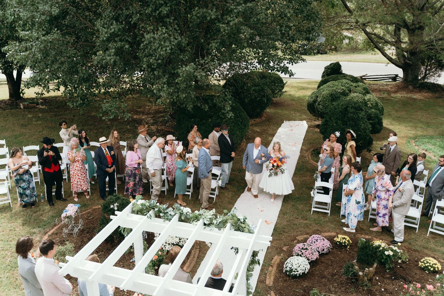 Wedding guests gather outdoors under trees for a garden ceremony with white chairs and floral decorations. East Coast wedding photographer