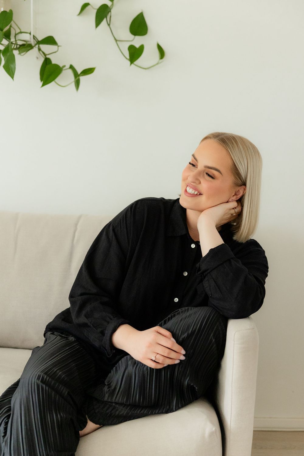 Person in black outfit lounges casually on white couch with green plants in background.