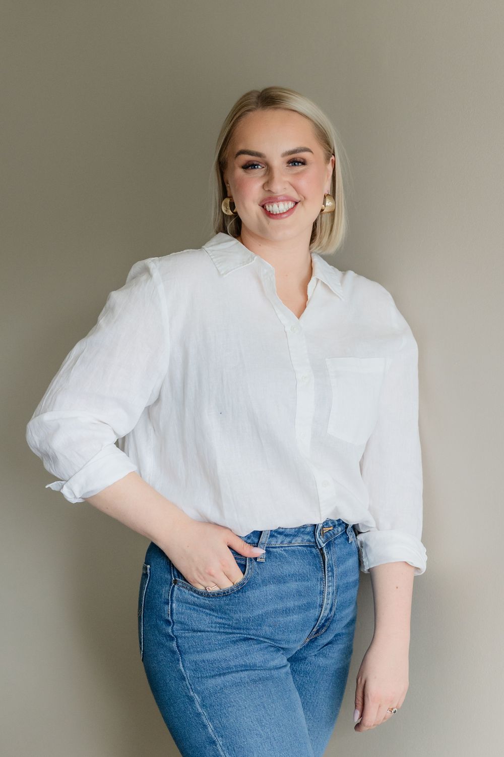 Person in white blouse and blue jeans poses against neutral wall in casual lifestyle photoshoot.