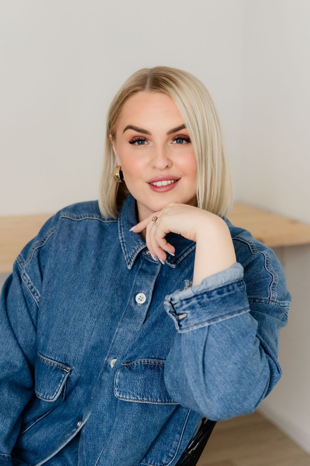 Stylish portrait of someone wearing oversized denim jacket while sitting against neutral background.