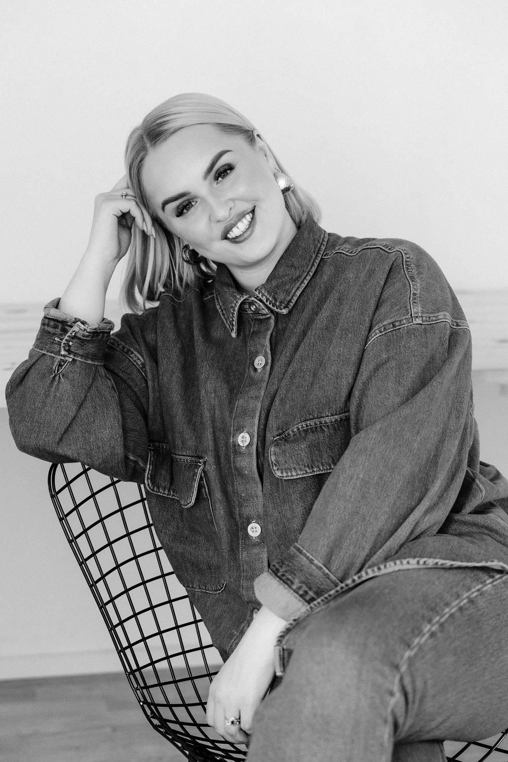 A black and white portrait of someone posing in denim on a wire chair with artistic lighting.