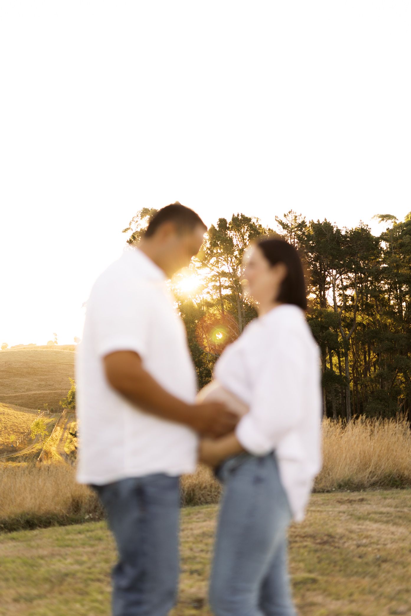 A couple dances together in a grassy field at sunset with golden light filtering through pine trees in the background.