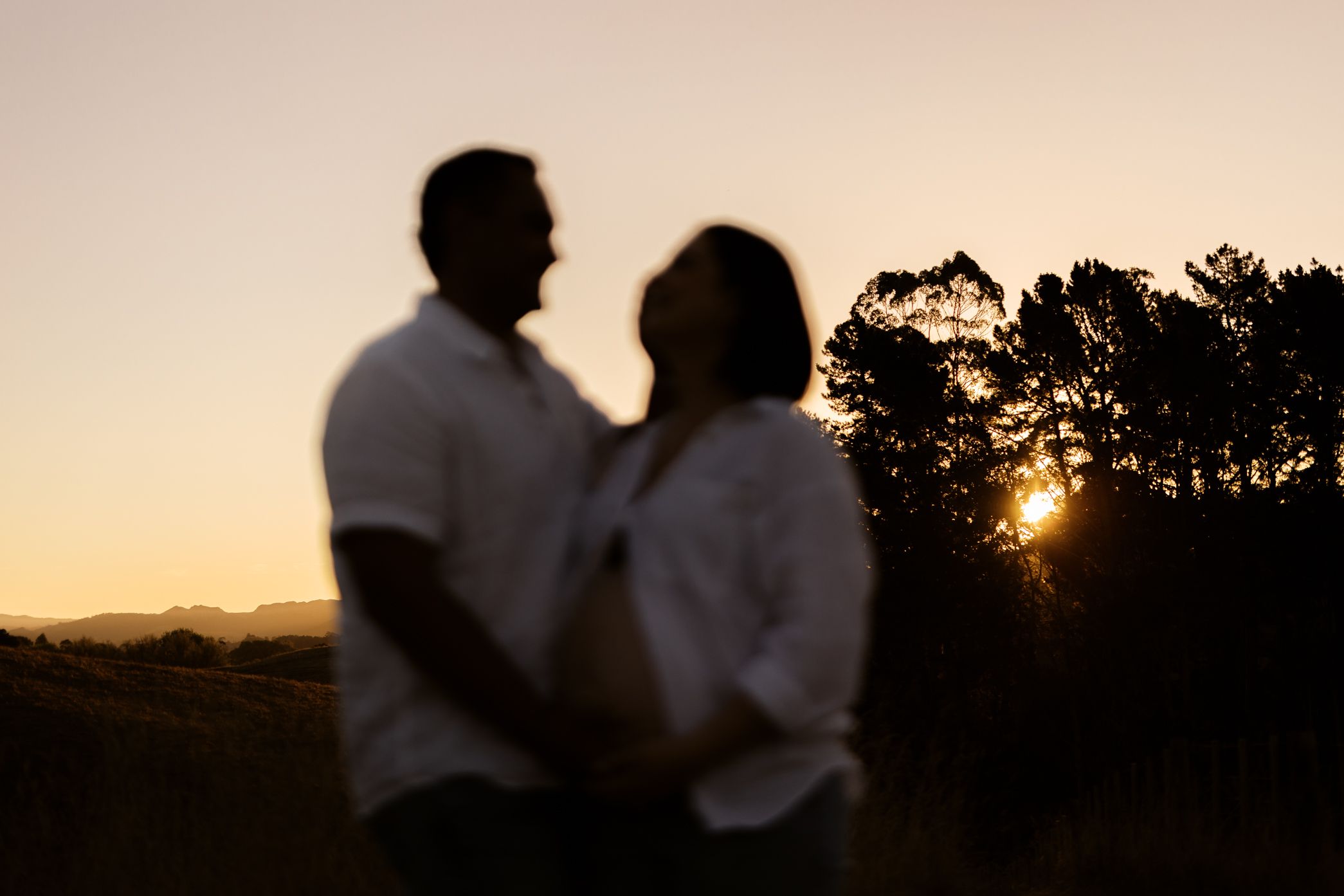 Silhouettes of a couple embrace during golden sunset with trees in the background.