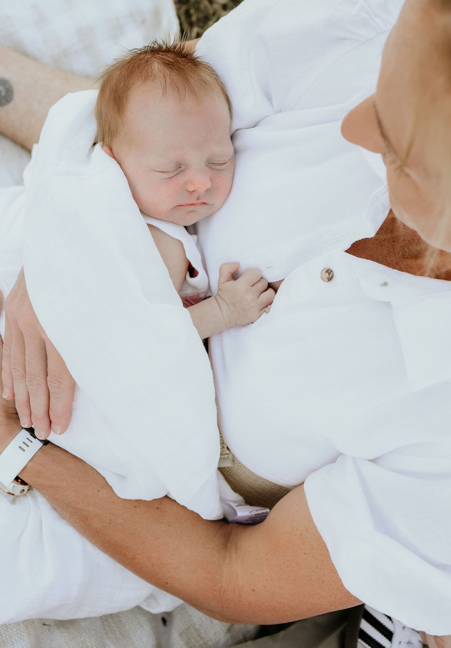 A sleeping newborn baby wrapped in soft white blankets being gently cradled by caring hands.