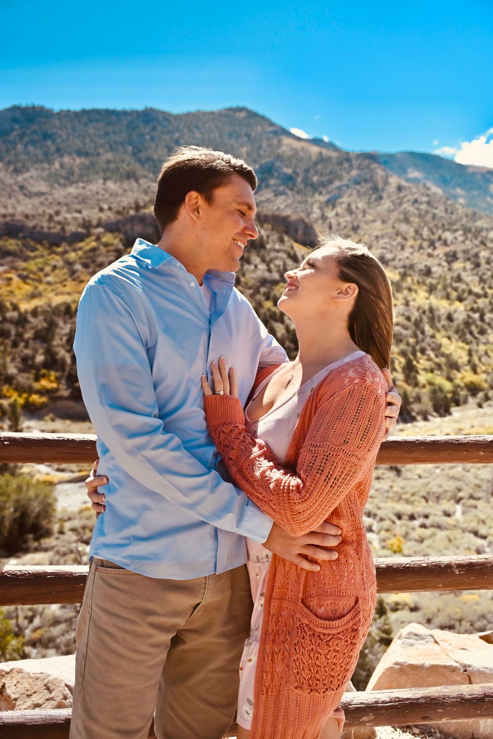 Joyful couple laughing and embracing on a wooden overlook with stunning mountain scenery behind them.