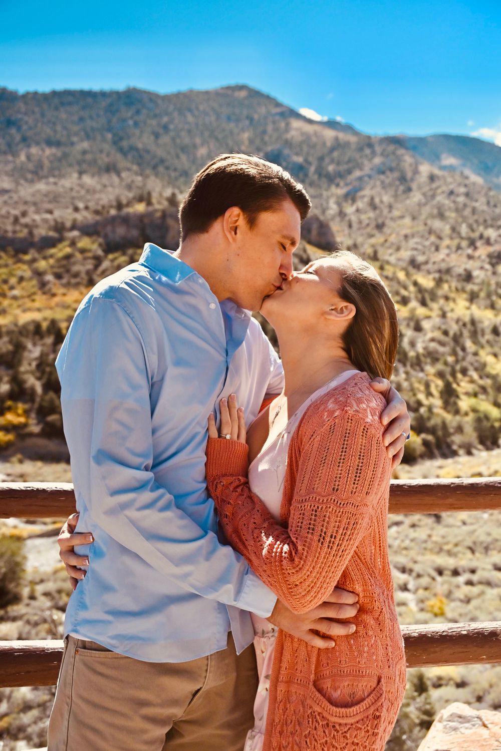 Couple sharing a kiss on a rustic wooden fence overlook with colorful mountain landscape in background.