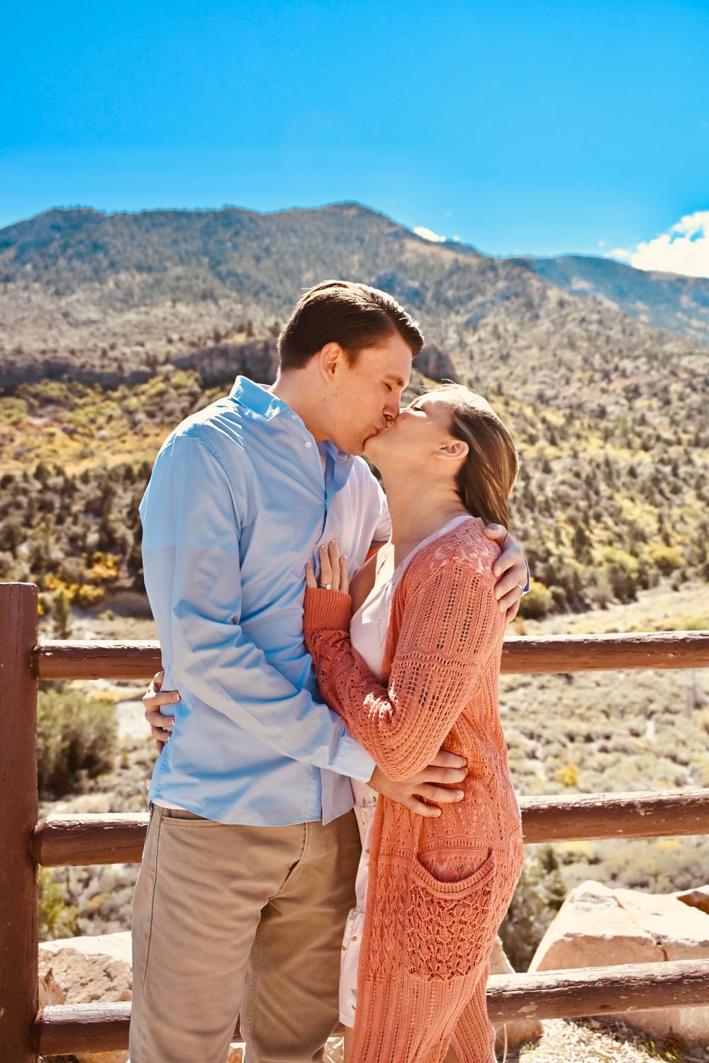 Romantic couple kissing on a wooden railed overlook surrounded by breathtaking mountain scenery.