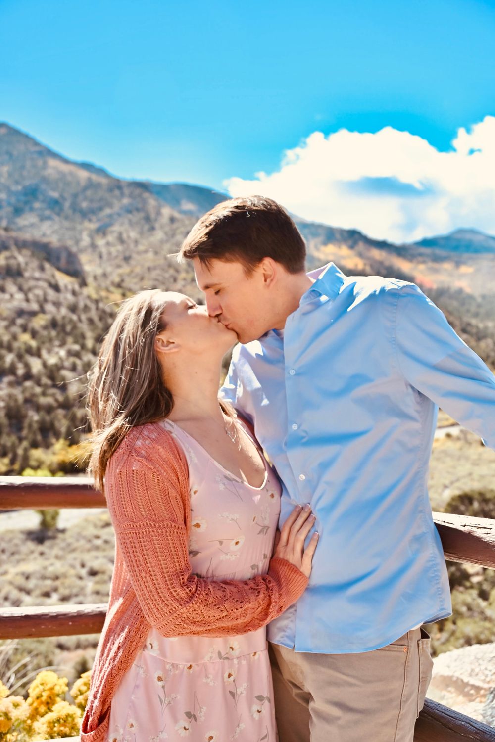 Couple sharing a tender kiss on a mountain overlook with blue sky and rugged terrain behind them.