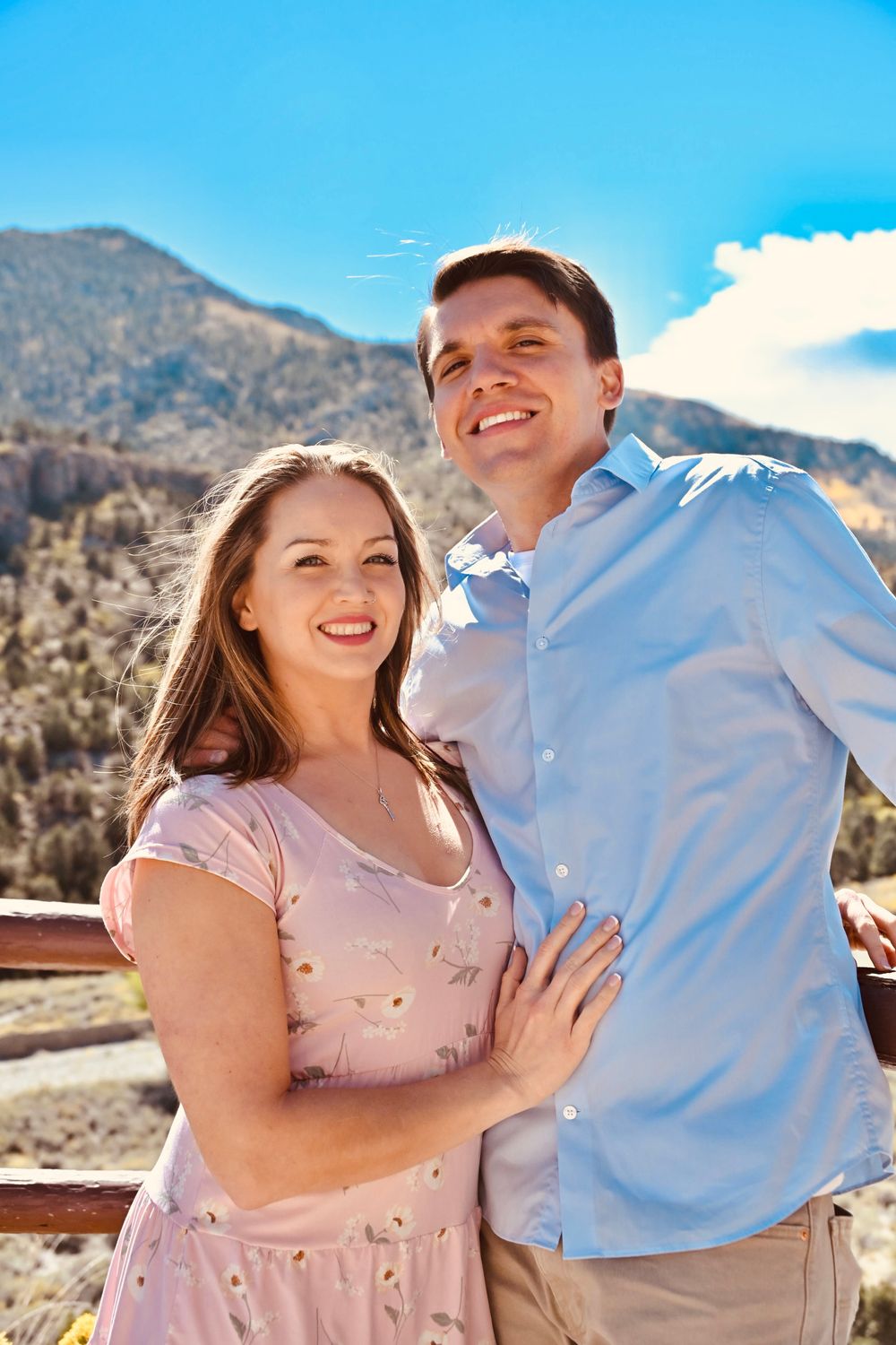 Smiling couple posing together at a mountain viewpoint, woman in floral pink dress and man in blue shirt.