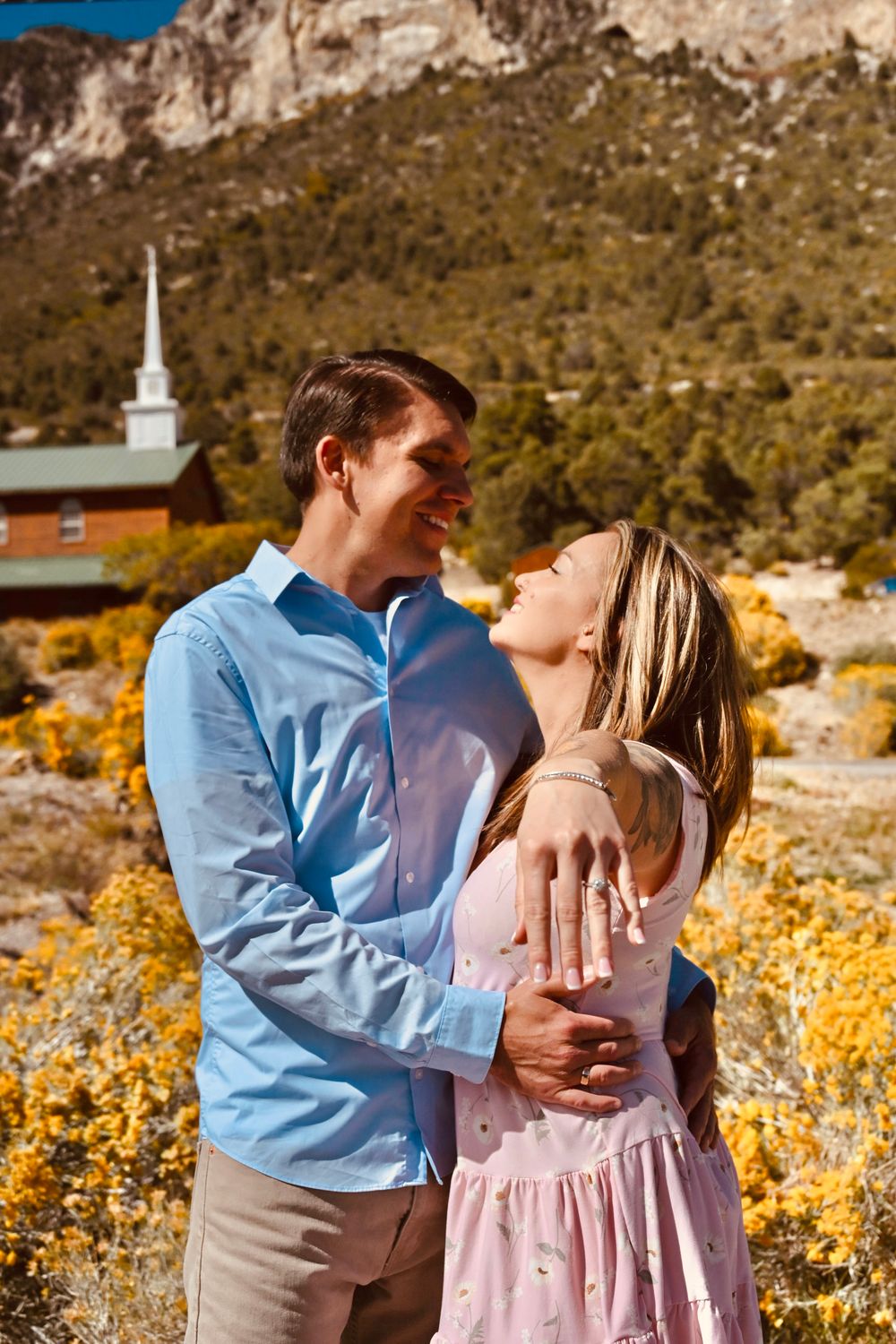 Couple embracing and smiling at each other near a white steeple church surrounded by autumn foliage.