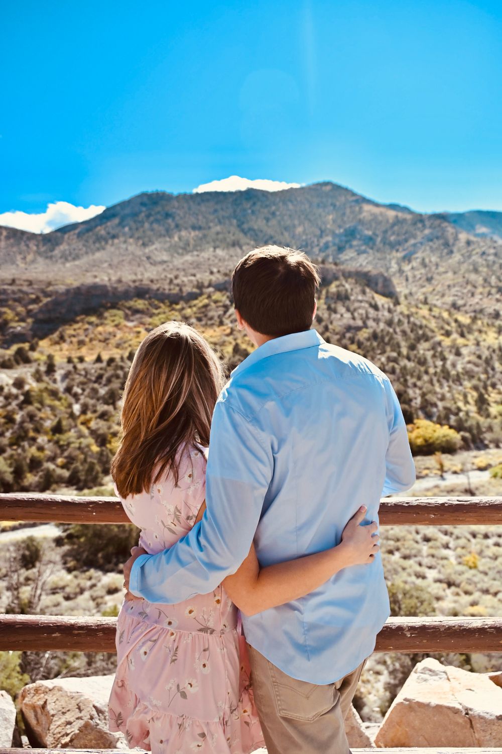 Couple standing arm in arm from behind, enjoying a stunning mountain landscape view on a clear day.