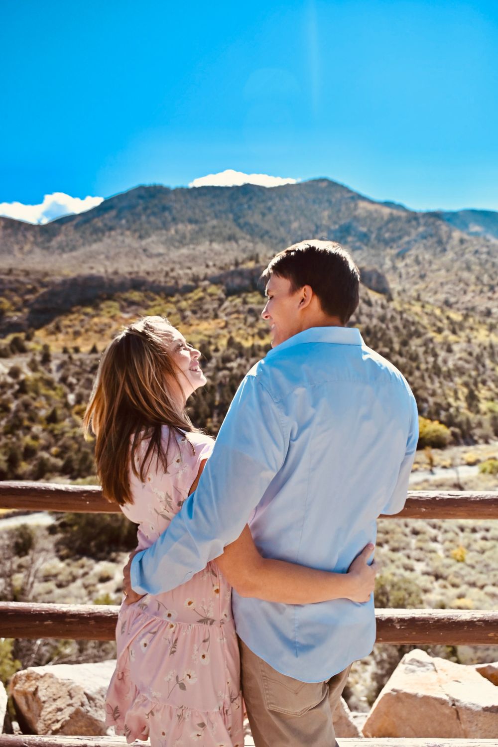 Couple laughing and embracing at a scenic mountain overlook with blue skies and rolling hills behind them.