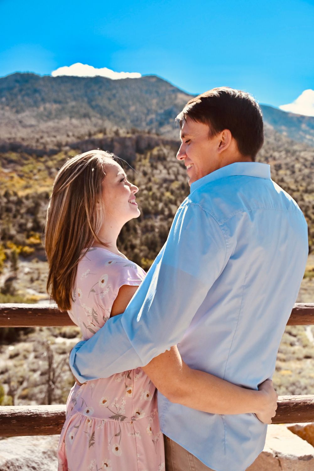 Couple holding each other close and smiling on a mountain overlook with a beautiful landscape backdrop.