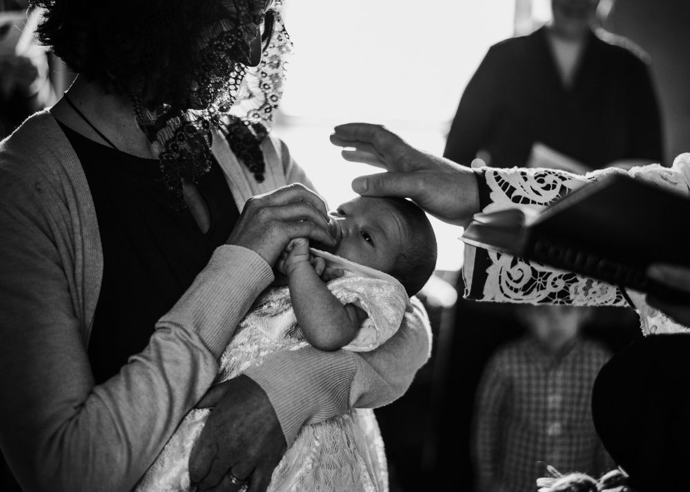At a Catholic Baptism, a woman in a mantilla veil is holding a newborn baby who is photographed being blessed by the priest.