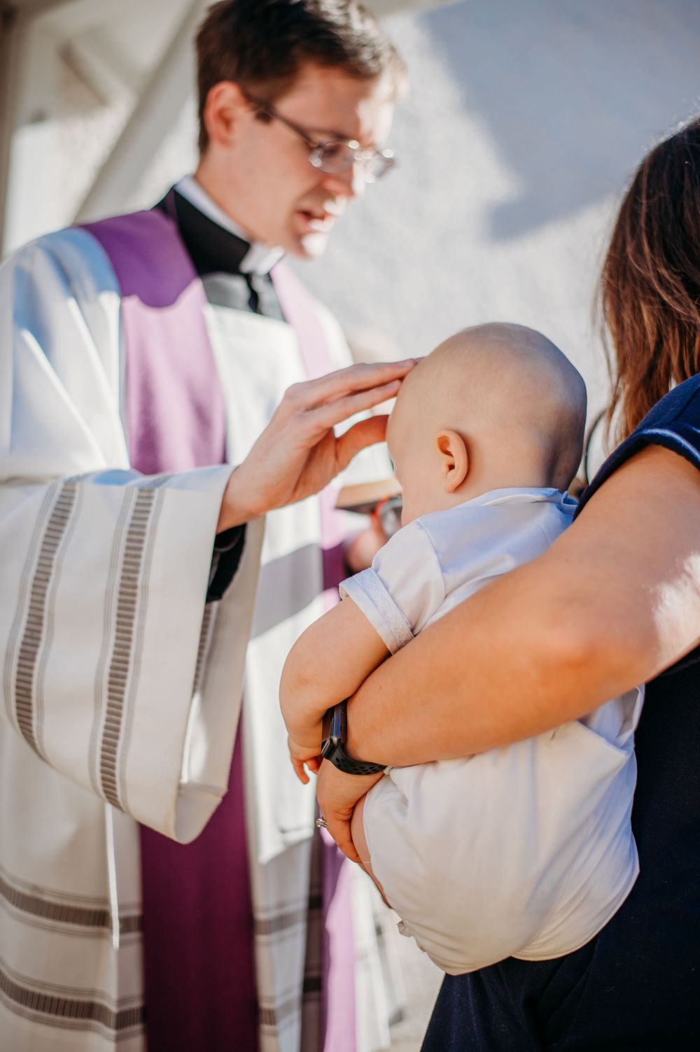 Outside of a small chapel, a young baby is being blessed by a Catholic priest before his Baptism, documented by an event photographer.