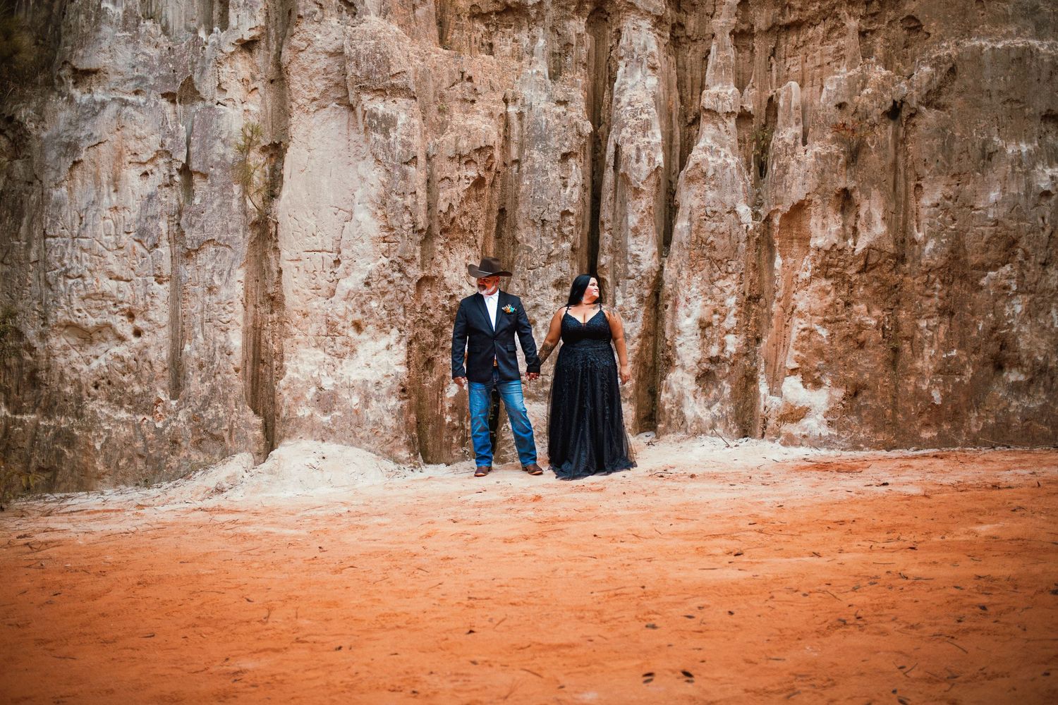 A couple stands against a rustic stone wall backdrop in an outdoor setting.