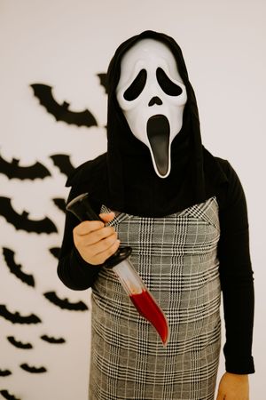 Person in Ghostface mask from Scream holding bloody knife against backdrop of black paper bats.