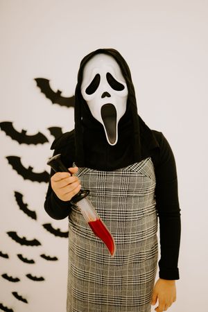 Figure wearing Scream mask and plaid dress poses with knife against wall with bat decorations.