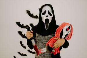 Person in Ghostface costume holds knife and mask against white wall with black bats.