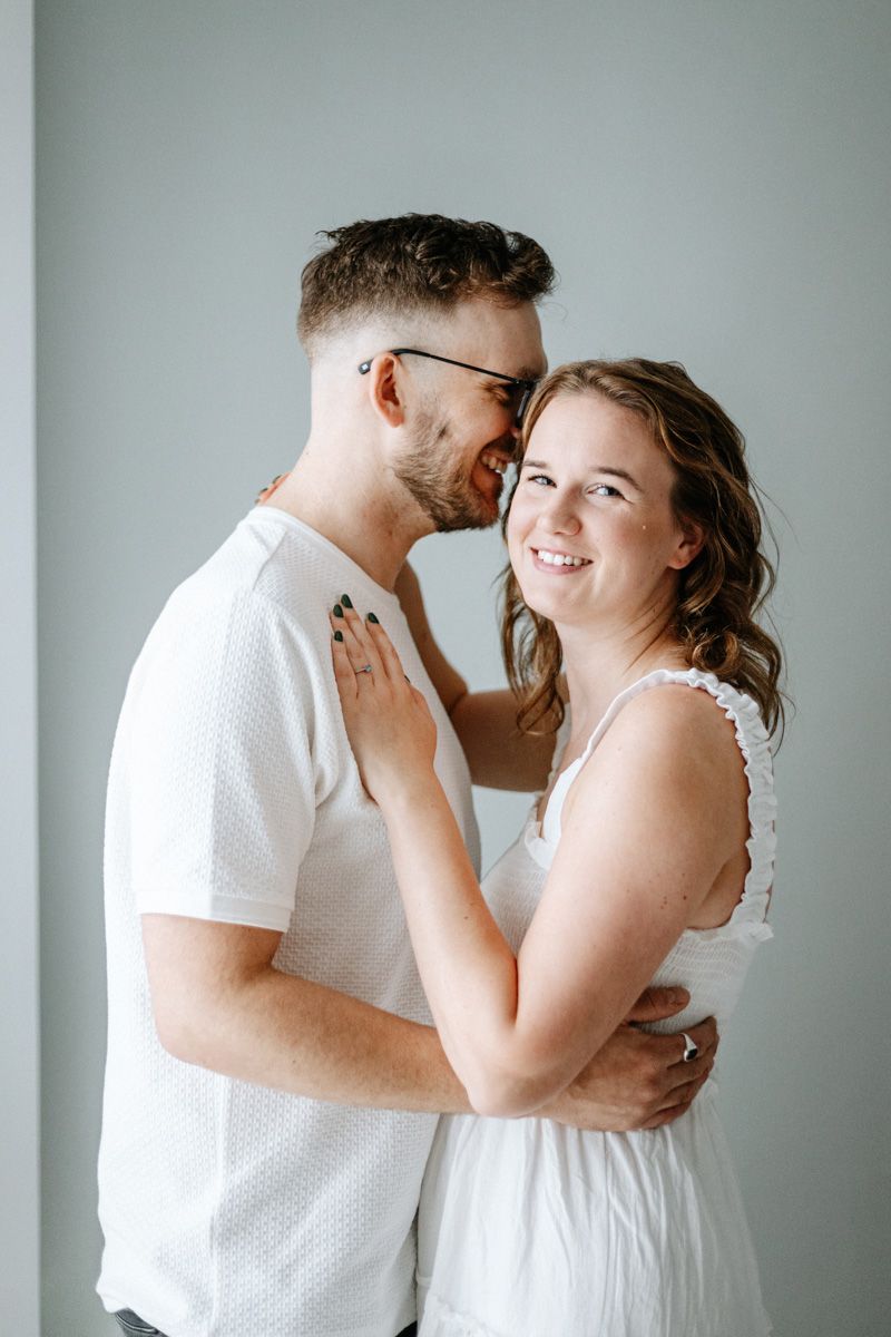 A romantic couple in white outfits share an intimate moment while embracing against a light gray background.