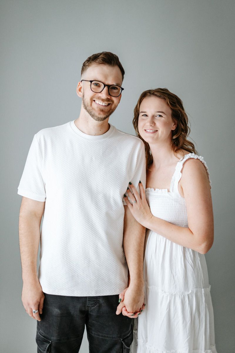 A couple wearing white tops poses together against a gray background in a minimalist studio setting.